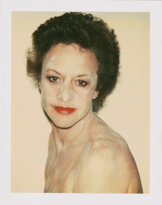 The image depicts a person's upper body, including the head, neck, and shoulders. The individual is facing slightly to the side with their head turned towards the camera. They have distinct facial features, fair complexion, prominent eyebrows, and bright red lipstick. Their dark hair is styled in a voluminous, slightly wavy manner. The person appears unclothed on the shoulders, which are visible in the image. The background is plain and of a neutral, light tone. No jewelry or objects are visible.