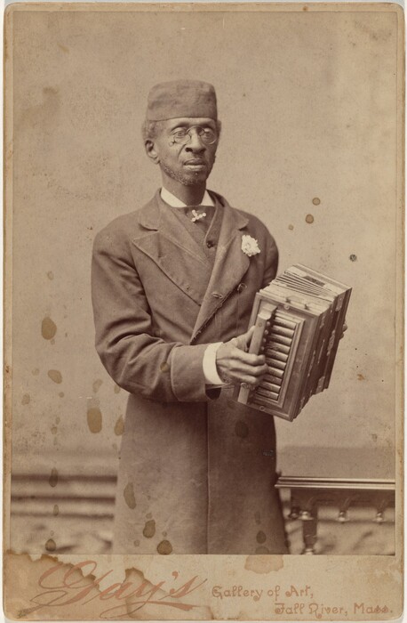 The image shows a man from the waist up in a standing position. He is facing slightly to the right and holding a musical instrument, possibly a concertina, in both hands. His facial features include a neatly trimmed beard and mustache, round glasses, short gray hair, and a distinguished expression. He is dressed in a double-breasted coat over a high-collared shirt, with a light-colored flower pinned to his left lapel. He is wearing a tall, rounded hat that matches the tone of his coat. The background is plain with some visible texture and a simple rail or banister is visible at the bottom right, suggesting an interior space. The photo shows signs of age.