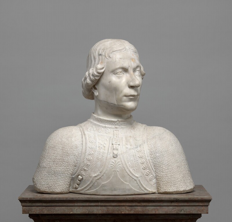 The head and shoulders of a cleanshaven man are carved from white marble lightly streaked in gray in this freestanding sculpture. In this view, the shoulders are square to us, but he turns his head to our right. His left eyebrow, to our right, appears to cocked as he looks into the distance, and he has a sweeping nose with a rounded tip. His long upper lip overhangs the lower lip in a slight overbite. His hair is parted down the middle and ends in curls that angle from eye-level framing his forehead to ear-level at the back of his neck. He wears a high-collared breastplate over linked mail. The breastplate is ornamented with bands of flowers, dots, and other geometric designs. A strap and buckle hang from the neck area. The sculpture ends in a blunt, straight edge across the bottom, and it rests on a brown stone pedestal in front of a gray background.
