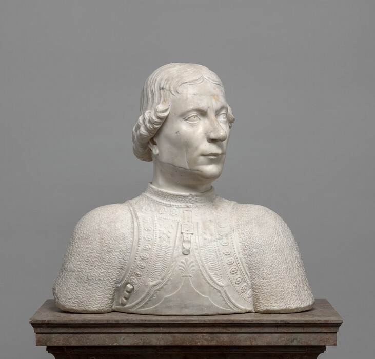 The head and shoulders of a cleanshaven man are carved from white marble lightly streaked in gray in this freestanding sculpture. In this view, the shoulders are square to us, but he turns his head to our right. His left eyebrow, to our right, appears to cocked as he looks into the distance, and he has a sweeping nose with a rounded tip. His long upper lip overhangs the lower lip in a slight overbite. His hair is parted down the middle and ends in curls that angle from eye-level framing his forehead to ear-level at the back of his neck. He wears a high-collared breastplate over linked mail. The breastplate is ornamented with bands of flowers, dots, and other geometric designs. A strap and buckle hang from the neck area. The sculpture ends in a blunt, straight edge across the bottom, and it rests on a brown stone pedestal in front of a gray background.