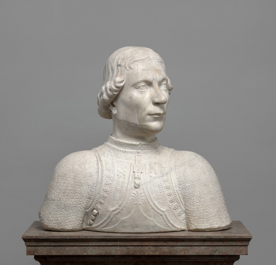The head and shoulders of a cleanshaven man are carved from white marble lightly streaked in gray in this freestanding sculpture. In this view, the shoulders are square to us, but he turns his head to our right. His left eyebrow, to our right, appears to cocked as he looks into the distance, and he has a sweeping nose with a rounded tip. His long upper lip overhangs the lower lip in a slight overbite. His hair is parted down the middle and ends in curls that angle from eye-level framing his forehead to ear-level at the back of his neck. He wears a high-collared breastplate over linked mail. The breastplate is ornamented with bands of flowers, dots, and other geometric designs. A strap and buckle hang from the neck area. The sculpture ends in a blunt, straight edge across the bottom, and it rests on a brown stone pedestal in front of a gray background.