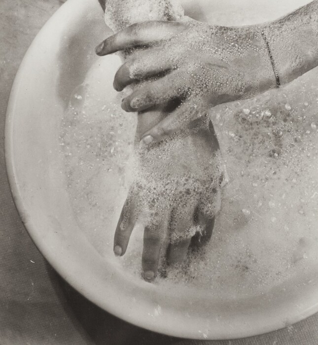 This is a photograph of hands being washed in a basin filled with soapy water. The image shows a pair of hands submerged in frothy, bubble-laden water contained in a round basin. The photograph is in black and white, highlighting the textures and contrasts between the smooth skin of the hands and the effervescent bubbles. The focus is on the way the soap suds cling to the skin, and the way the light interacts with the bubbles. The close-up perspective allows for a detailed view of the washing process, emphasizing cleanliness and the tactile sensation of water and soap.