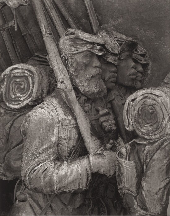 The sculpture depicts a group of soldiers marching in close formation. The figures wear caps and uniforms with coats and carry rolled blankets or bags over their shoulders. They appear to be holding rifles, suggesting they are infantrymen. The sculpture is crafted from a dark material, possibly bronze, with a weathered patina. The texture is rough, enhancing the realistic depiction of clothing and facial hair. The figures are carved in high relief, giving them a three-dimensional look. The expression and detail suggest attention to realistic features typical of soldiers.