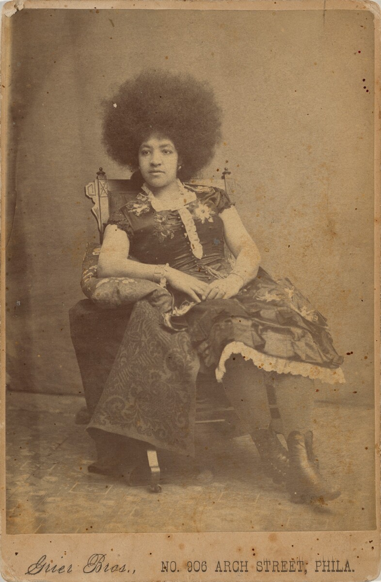 The image shows a woman seated in a three-quarter length portrait. She is sitting in a relaxed position on a decorative chair, with her right arm resting on a small cushion on the armrest. Her left arm lies across her lap. The woman has a voluminous afro hairstyle and a composed expression. She wears a dress with floral patterns, lace details, and high lace-up boots. She is adorned with a bracelet on her right wrist. The backdrop is a textured surface, with the caption "Grier Bros., No. 906 Arch Street, Phila." at the bottom of the photograph.