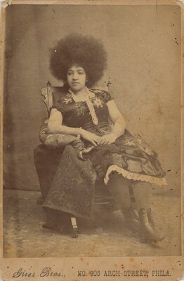 The image shows a woman seated in a three-quarter length portrait. She is sitting in a relaxed position on a decorative chair, with her right arm resting on a small cushion on the armrest. Her left arm lies across her lap. The woman has a voluminous afro hairstyle and a composed expression. She wears a dress with floral patterns, lace details, and high lace-up boots. She is adorned with a bracelet on her right wrist. The backdrop is a textured surface, with the caption "Grier Bros., No. 906 Arch Street, Phila." at the bottom of the photograph.