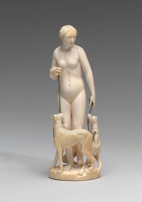 The sculpture depicts a standing nude female figure holding chains attached to two dogs. The woman is looking downwards towards one of the dogs. The sculpture is made from a smooth, ivory-colored material with a polished surface. Relative to us, the sculpture is small and detailed, capturing intricate elements such as the woman's facial features, hair, and the muscles and posture of the dogs around the woman's feet.