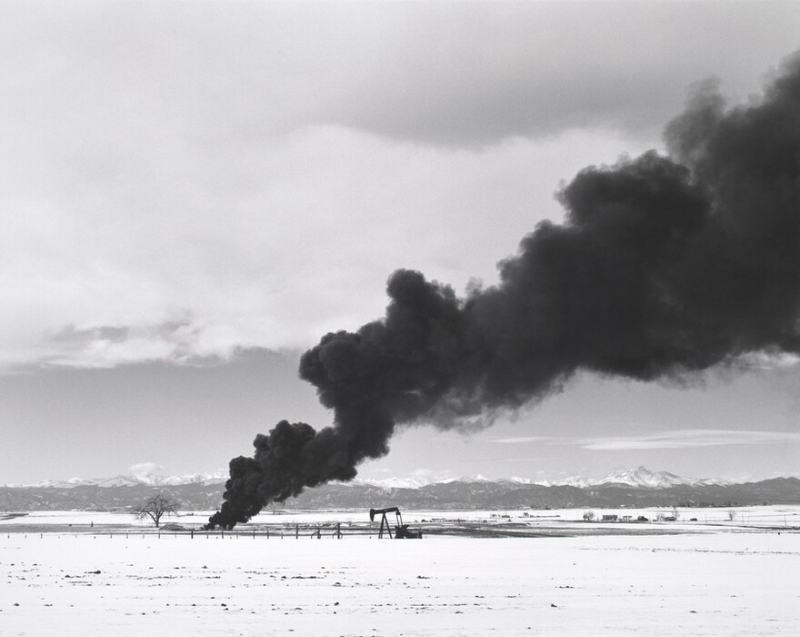 The image shows a landscape with mountains in the distance, a leafless tree on the left, and an oil pump jack on the right. The sky is clouded with dark smoke, contrasting with the snowy ground in the foreground. The scene is in black, white, and gray colors.
