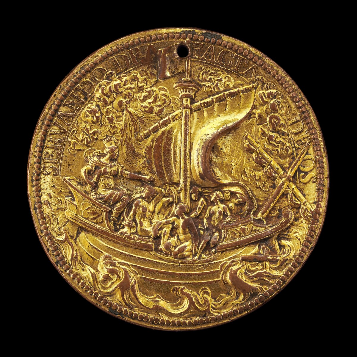 A photograph of a gold-colored medallion is shown. The medallion depicts a ship with a sail surrounded by waves and clouds. Figures on the ship are engaged in activities. The relief work is detailed, particularly in the figures and sail. An inscription in a classical style frames the scene on the edge of the medallion. The craftsmanship suggests a historical or commemorative purpose, highlighting fine artistry and detailed representation.