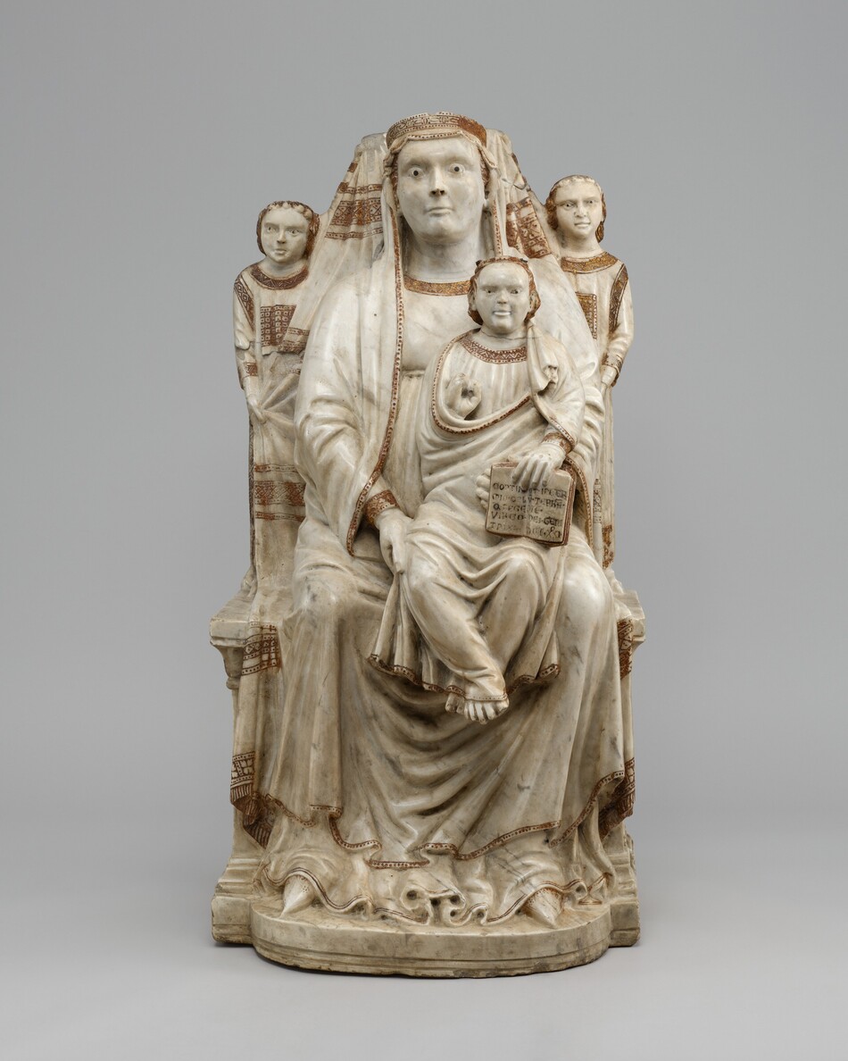 A woman sitting on a throne with a child on her lap and two angels are carved from white marble and decorated with a few areas of gold in this freestanding sculpture. All four people face us in this photograph, and they have round cheeks, square jawlines, and closed lips. Black dots are painted for pupils but without irises or other paint on their faces, they all look startled or intense. The woman wears a gold crown over a cloth that drapes over her head, shoulders, and across her lap. Her knees are apart, and pointed shoes peek out under the rippling hem of her dress. She braces the child’s torso with her left hand, to our right, and touches his upper leg with the other hand. The child’s adult-like face is framed by gold hair. He holds up one hand, though the fingers have been broken off, and holds an open book facing out propped on his leg with his other hand. Writing on the pages has been painted. Two child-like, wingless angels stand on the arms of the throne behind the woman’s shoulders and hold a cloth up behind her head. On all four people, their hair and the necklines, hems, and cuffs of their garments have been painted with gold geometric designs. The Latin text on the book reads, "CONTINET IN GERMIO CELV TERRA Q REGENTE VIRGO DEI GENETRIX MCCCXXI.”