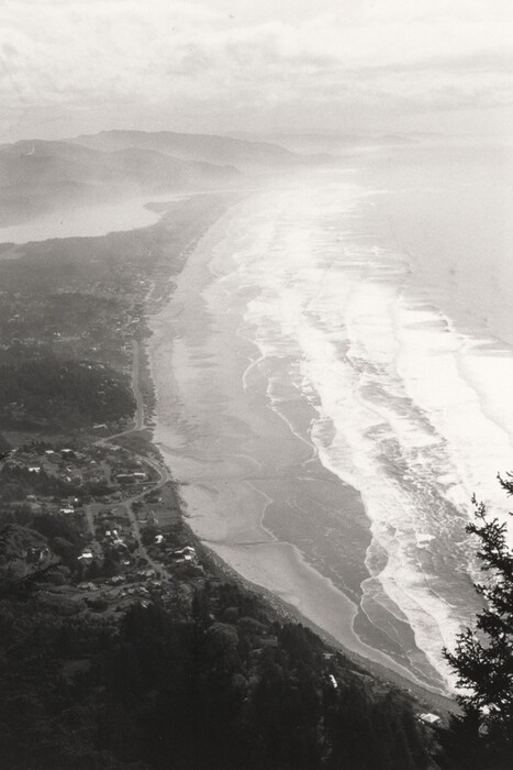The image shows an aerial view of a coastal landscape. There is a forested area near the edge, small settlements along a winding shore road, and a low horizon. The sky dominates the upper part with gray clouds. Mountain ridges are visible in the background, blending into the sky. The color palette is black and white, giving a timeless quality. Waves flow along the shoreline with white crests contrasting the water.