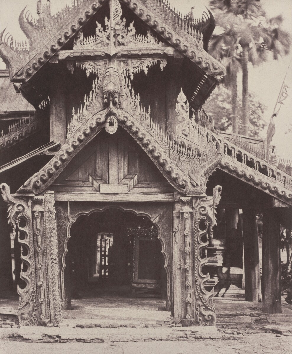 This is a photograph of a wooden building with intricate architectural details. The gable end is adorned with swirling patterns and decorative elements. The entrance has a slightly arched doorway surrounded by detailed woodwork, with a carved panel above it. The roof edges have decorative trim and flourishes. In the background are tall palm trees and foliage. The craftsmanship of the building showcases attention to detail and artistry, capturing a moment of cultural heritage in wooden architecture.