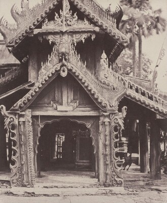 This is a photograph of a wooden building with intricate architectural details. The gable end is adorned with swirling patterns and decorative elements. The entrance has a slightly arched doorway surrounded by detailed woodwork, with a carved panel above it. The roof edges have decorative trim and flourishes. In the background are tall palm trees and foliage. The craftsmanship of the building showcases attention to detail and artistry, capturing a moment of cultural heritage in wooden architecture.