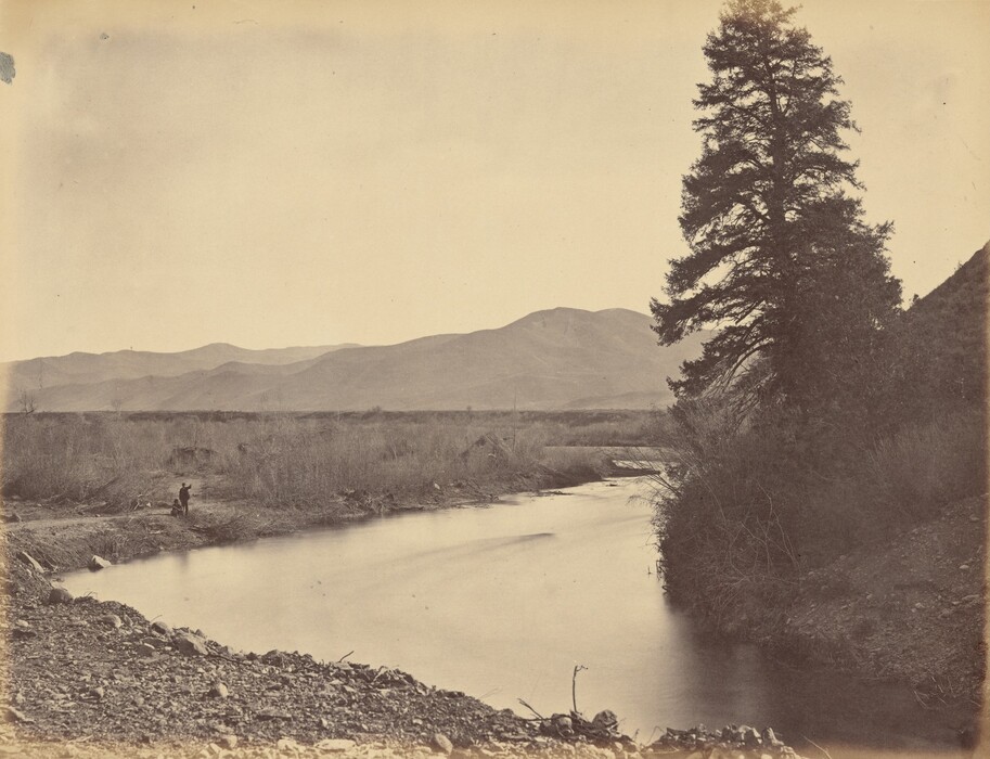 The image shows a landscape with a river flowing through it. Rocky ground lines the left riverbank where a person is sitting or crouching. A large tree is on the right, with mountains in the distance. The sky is expansive above. The color scheme is sepia, giving an antique feel. The image is a photograph capturing details like water ripples and tree foliage.