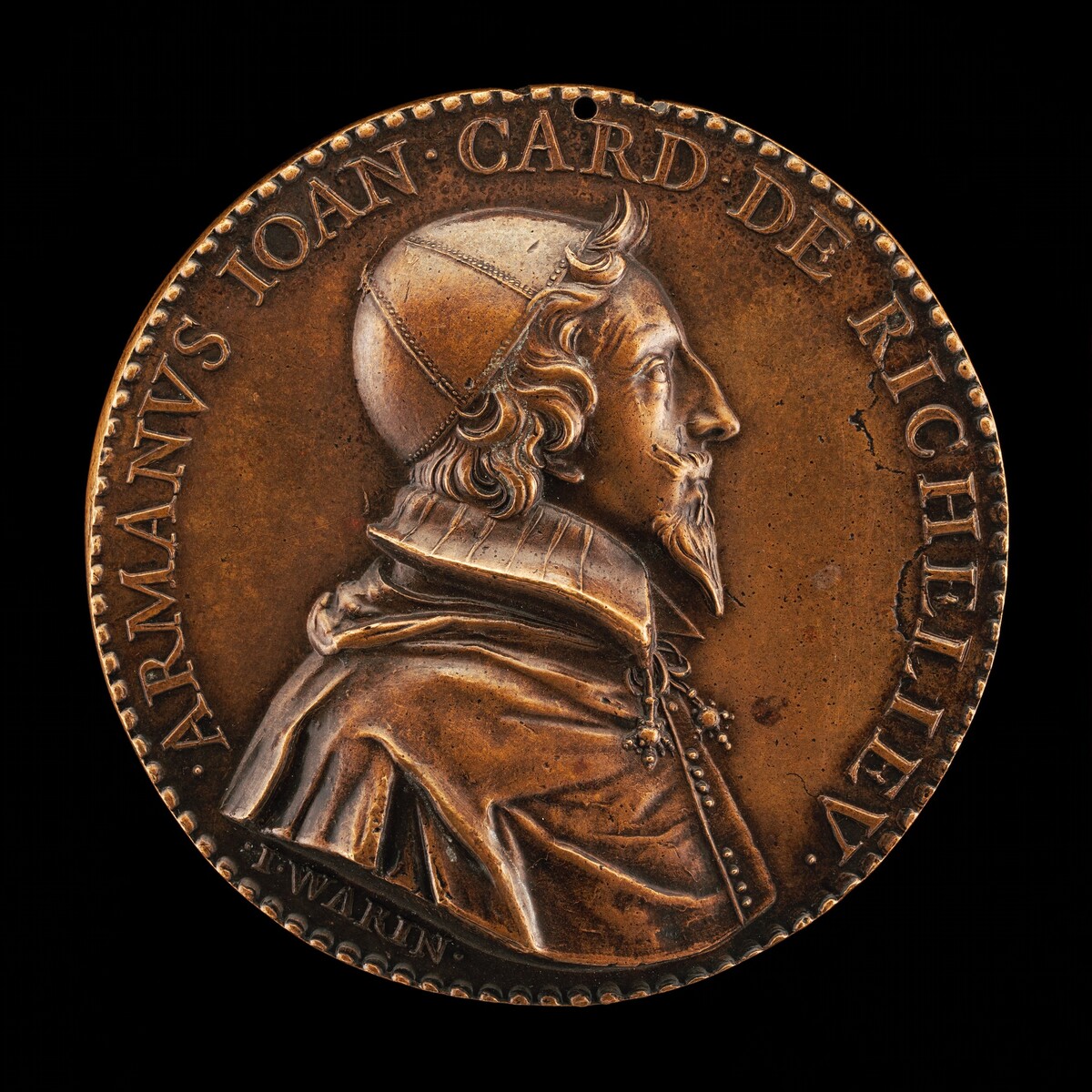 The sculpture is a bas-relief medallion featuring a profile view of a man identified as "ARMANVS IOAN CARD DE RICHELIEV." The man is depicted wearing a cap and an elaborate, high-collared garment with visible details in the fabric and folds, along with ornamental chains and a pendant. The material of the medallion appears to be a warm, bronze metal. The sculpting accurately captures facial features such as the man's wavy hair, beard, and prominent nose. The round medallion includes the artist's name, "J. WARIN," engraved along the bottom edge. It is designed to have a small hole at the top.