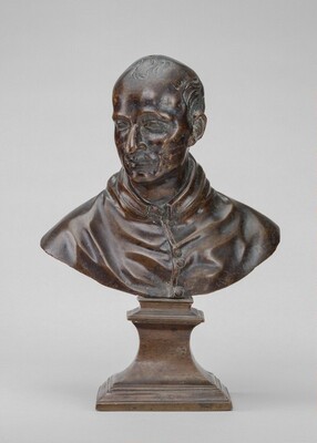 The sculpture is a bust of a mature man with a thoughtful expression and neatly combed hair. It is made from dark brown material, likely polished wood. The details of the man's face and clothing, such as buttons on his garment and folds around the neck area, are finely carved. The bust is mounted on a geometric base, evoking a sense of classical elegance.