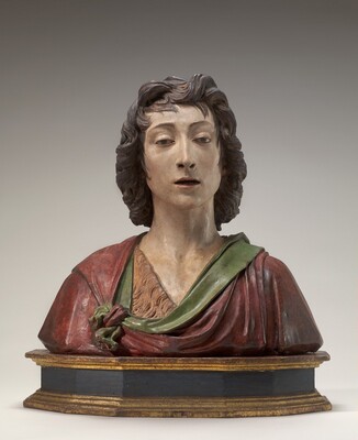 The sculpture is a detailed bust of a young man with pale skin and wavy brown hair that almost reaches to his shoulders, seen from the chest up. He has thin, arched eyebrows, a long, straight nose, and thin red lips that are partially open. A dark red and green cloth or cloak is draped over his shoulders and tied near his left arm, and it appears to cover a light brown garment with the texture of fur. The sculpture has no base.