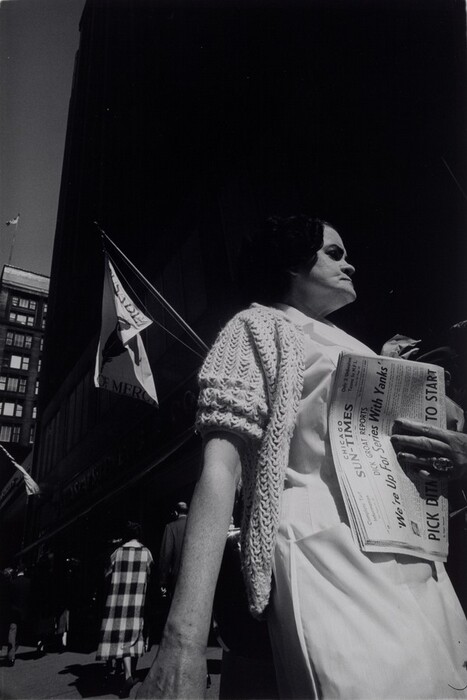 The image shows a woman captured from a low angle, with the upper half of her body in view. She is standing upright, facing slightly to the right of the frame. Her facial features include prominent cheekbones, and she has short, dark hair. She is wearing a light-colored dress with a crochet cardigan over it. In her right hand, she holds a folded newspaper labeled "CHICAGO SUN-TIMES." She is also wearing a ring on her right hand, featuring a large stone. The background includes the exterior of a multi-story building, partially visible, with a flag visible on it, and bustling street scenes beneath with pedestrians walking in various directions. A flag displaying a bird and the word "MERC" is visible, and the sky is clear, suggesting daytime.