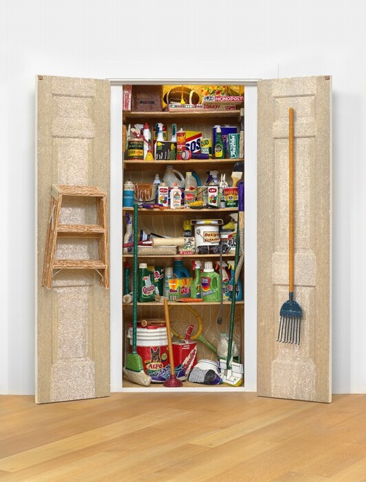 The sculpture depicts a storage closet with open doors revealing various household items inside. The doors are white, and a step stool hangs on the left one while a broom hangs on the right. The shelves are filled with typical household products: mostly cleaning supplies, painting supplies, and other various objects, like a saw, a plunger, a glue gun, and a flashlight. Upon closer inspection, it is revealed that everything in the closet, including the doors, has been decorated with tiny, colorful beads. The beaded closet is set against white walls and a wooden floor.