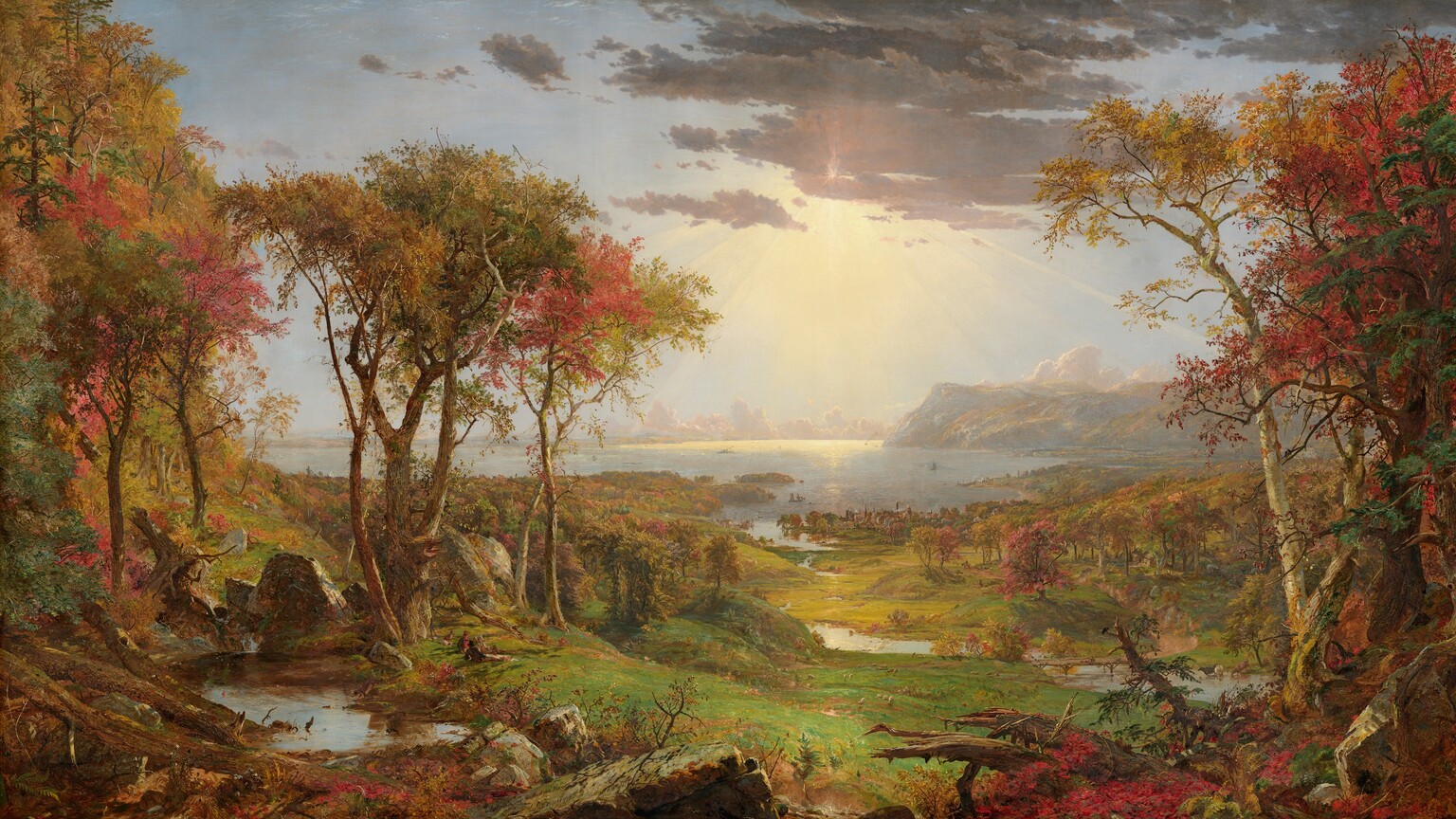 We look out onto a sweeping, panoramic view with trees, their leaves fiery orange and red, framing a view of a distant body of water under a sun-streaked sky in this long, horizontal landscape painting. The horizon comes about halfway up the composition, and is lined with hazy mountains and clouds in the deep distance. Close examination slowly reveals minuscule birds tucked into the crimson-red, golden yellow, and deep, sage-green leaves of the trees to either side of the painting. Closest to us, vine-covered, fallen tree trunks and mossy gray boulders line the bottom edge of the canvas. Beyond a trickling waterfall and small pool near the lower left corner, and tiny within the scale of the landscape, a group of three men and their dogs sit and recline around a blanket and a picnic basket, their rifles leaning against a tree nearby. The land sweeps down to a grassy meadow crossed by a meandering stream that winds into the distance, at the center of the painting. Touches of white and gray represent a flock of grazing sheep in the meadow. A low wooden bridge spans the stream to our right, and a few cows drink from the riverbank. Smoke rises from chimneys in a town lining the riverbank and shoreline beyond, and tiny white sails and steamboats dot the waterway. Light pours onto the scene with rays like a starburst from behind a lavender-gray cloud covering the sun, low in the sky. The artist signed the painting as if he had inscribed the flat top of a rock at the lower center of the landscape with his name, the title of the painting, and date: “Autumn – on the Hudson River, J.F Cropsey, London 1860.”