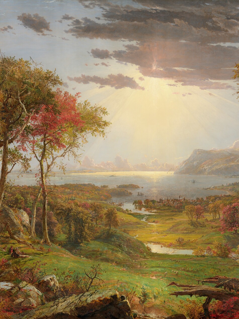 We look out onto a sweeping, panoramic view with trees, their leaves fiery orange and red, framing a view of a distant body of water under a sun-streaked sky in this long, horizontal landscape painting. The horizon comes about halfway up the composition, and is lined with hazy mountains and clouds in the deep distance. Close examination slowly reveals minuscule birds tucked into the crimson-red, golden yellow, and deep, sage-green leaves of the trees to either side of the painting. Closest to us, vine-covered, fallen tree trunks and mossy gray boulders line the bottom edge of the canvas. Beyond a trickling waterfall and small pool near the lower left corner, and tiny within the scale of the landscape, a group of three men and their dogs sit and recline around a blanket and a picnic basket, their rifles leaning against a tree nearby. The land sweeps down to a grassy meadow crossed by a meandering stream that winds into the distance, at the center of the painting. Touches of white and gray represent a flock of grazing sheep in the meadow. A low wooden bridge spans the stream to our right, and a few cows drink from the riverbank. Smoke rises from chimneys in a town lining the riverbank and shoreline beyond, and tiny white sails and steamboats dot the waterway. Light pours onto the scene with rays like a starburst from behind a lavender-gray cloud covering the sun, low in the sky. The artist signed the painting as if he had inscribed the flat top of a rock at the lower center of the landscape with his name, the title of the painting, and date: “Autumn – on the Hudson River, J.F Cropsey, London 1860.”