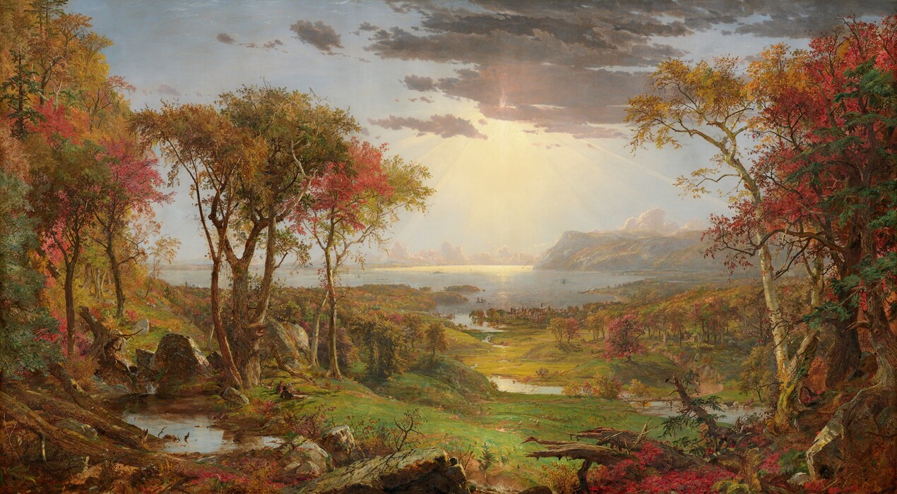 We look out onto a sweeping, panoramic view with trees, their leaves fiery orange and red, framing a view of a distant body of water under a sun-streaked sky in this long, horizontal landscape painting. The horizon comes about halfway up the composition, and is lined with hazy mountains and clouds in the deep distance. Close examination slowly reveals minuscule birds tucked into the crimson-red, golden yellow, and deep, sage-green leaves of the trees to either side of the painting. Closest to us, vine-covered, fallen tree trunks and mossy gray boulders line the bottom edge of the canvas. Beyond a trickling waterfall and small pool near the lower left corner, and tiny within the scale of the landscape, a group of three men and their dogs sit and recline around a blanket and a picnic basket, their rifles leaning against a tree nearby. The land sweeps down to a grassy meadow crossed by a meandering stream that winds into the distance, at the center of the painting. Touches of white and gray represent a flock of grazing sheep in the meadow. A low wooden bridge spans the stream to our right, and a few cows drink from the riverbank. Smoke rises from chimneys in a town lining the riverbank and shoreline beyond, and tiny white sails and steamboats dot the waterway. Light pours onto the scene with rays like a starburst from behind a lavender-gray cloud covering the sun, low in the sky. The artist signed the painting as if he had inscribed the flat top of a rock at the lower center of the landscape with his name, the title of the painting, and date: “Autumn – on the Hudson River, J.F Cropsey, London 1860.”