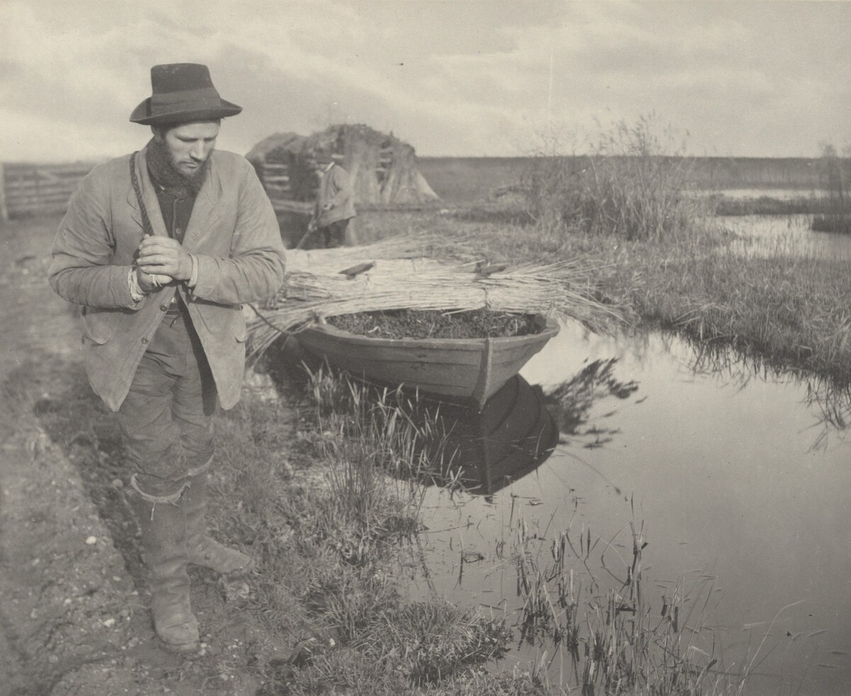 This is a photograph of a rural scene by a small waterway. The image captures an outdoor setting alongside a narrow water channel, with reflections of surrounding grass and a docked wooden boat floating in the water. The boat is laden with bundles of vegetation. In the background, a man stands near a stack of collected grasses or hay. Several elements of rural life are apparent, such as the dirt path, wooden fence sections, and the rustic nature of the landscape. The sky is partly cloudy, casting an even light across the scene.