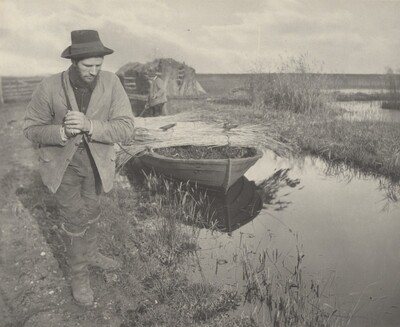 This is a photograph of a rural scene by a small waterway. The image captures an outdoor setting alongside a narrow water channel, with reflections of surrounding grass and a docked wooden boat floating in the water. The boat is laden with bundles of vegetation. In the background, a man stands near a stack of collected grasses or hay. Several elements of rural life are apparent, such as the dirt path, wooden fence sections, and the rustic nature of the landscape. The sky is partly cloudy, casting an even light across the scene.