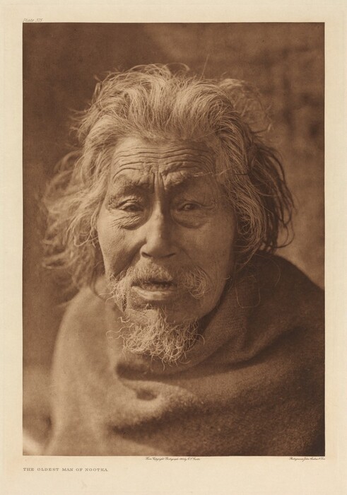 The image shows the upper half of an elderly man's body with a focus on his face. He appears to be seated, with his shoulders slightly angled toward the viewer. His facial features are deeply lined and expressive, with prominent wrinkles across his forehead and under his eyes. He has a full head of unruly, gray hair that frames his face and an equally gray beard and mustache. His facial expression is neutral, with his mouth slightly open. He is wrapped in a blanket or cloak, which covers most of his body, leaving only the top of his shoulders and face visible. The background is blurred and neutral.