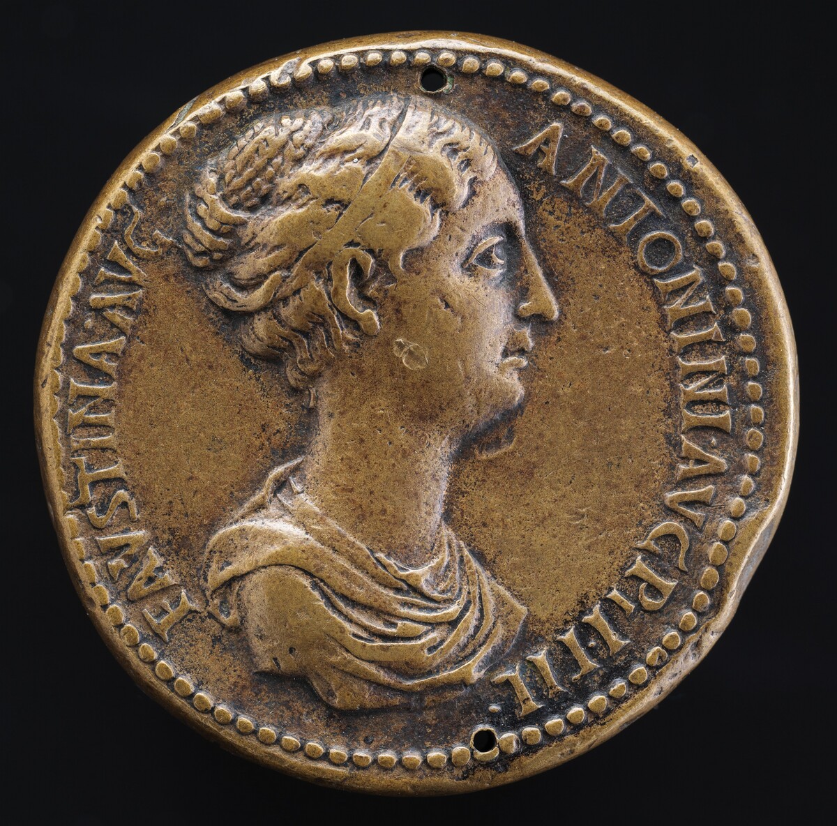 The image depicts a relief of a woman's profile facing right on an ancient Roman coin. The subject appears to be a classical figure, suggested by her attire and the style of the coin. The sculpture is made of a light brown material, possibly bronze. Text in Latin is inscribed along the border of the coin. The woman's hair is styled intricately and seems to be held up with a diadem or headband. There is a small hole near the edge of the coin, indicating it might have been used as a pendant or ornament. The texture of the coin shows signs of wear, suggesting its age and historical use.