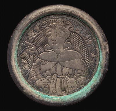 This is a photograph of a circular medallion or coin. The medallion features a raised relief depicting a robed figure with a halo holding a flower. The relief is set against a textured, diagonal crosshatch background. Surrounding the central figure are inscriptions that appear to form a name or title, including the letters "SA" and "LBERTVS." The item has a worn and weathered appearance, with a greenish patina along its rim.