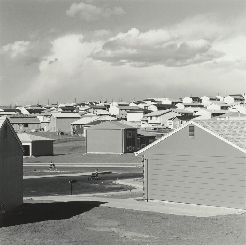A patchwork of peaked roofs spreads in front of us in this square black and white photograph. We look at the neighborhood from between two houses, where a child pushes a wagon or toy baby stroller in the driveway. Low, puffy clouds float in front of a hazier screen of clouds in the top half of the picture.