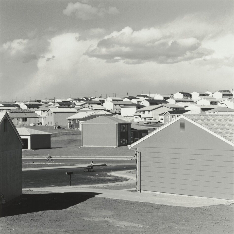 A patchwork of peaked roofs spreads in front of us in this square black and white photograph. We look at the neighborhood from between two houses, where a child pushes a wagon or toy baby stroller in the driveway. Low, puffy clouds float in front of a hazier screen of clouds in the top half of the picture.