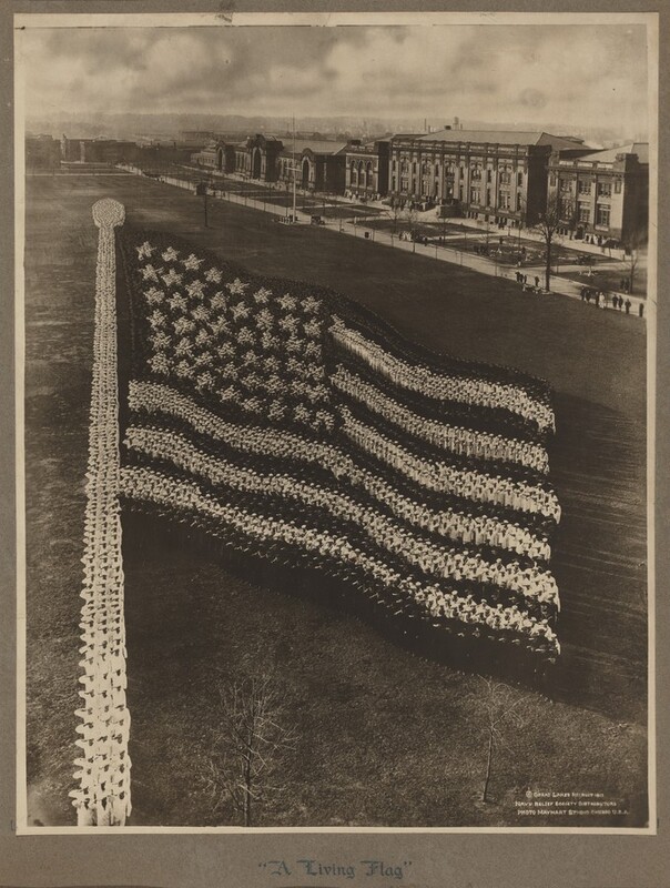 Hundreds of people wearing white or dark-colored uniforms stand in formation to make an American flag on a long, rectangular, grassy area in this black and white photograph. We can tell from the people closest to us that they stand at attention and salute. The flagpole is to our left, and the stars and stripes ripple downward as if the flag was lifted in a slight breeze. About a dozen people stand along the edge of the far side park, which is lined with four and five-story buildings. White clouds are out of focus in the sky above, which takes up about the top sixth of the photograph. Text printed in white against the dark photograph in the lower right corner begins with a copyright symbol and then reads, “Great Lakes Recruit 1917, Navy Relief Society Distributors, Photo Mayhart Studio Chicago U.S.A.” Text written in a calligraphic style on the gray mat under the photograph reads, “A Living Flag.”