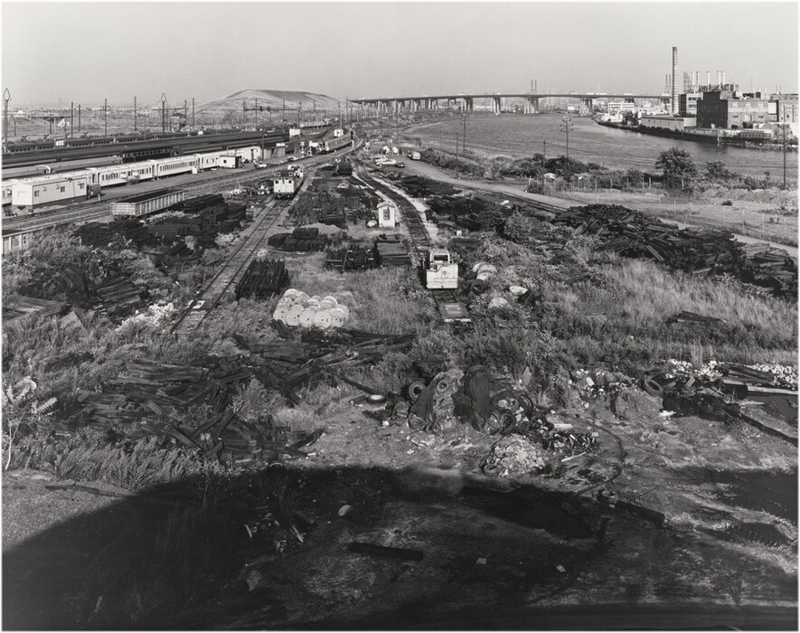The image depicts an elevated view of an industrial yard with tracks and scattered materials. The horizon is in the upper section of the image, featuring a flat expanse with a distant bridge. Train carriages line up parallel to the horizon, with industrial buildings and chimneys on the far right. Closest to us, there is industrial debris and materials scattered on the ground. The image is in black and white, emphasizing dark tones and textures. It is a photograph with a monochromatic color palette.