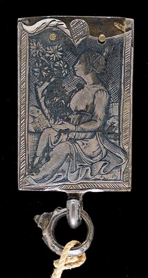 This is a photograph of an engraved metal object. The engraved image depicts a figure wearing a dress sitting on a chair, holding a bird, surrounded by floral elements. The figure is set against a patterned background with detailed engraving that enhances the texture of the garment and the foliage. The object's borders are adorned with a simple, repeating pattern, and there are metallic rivets visible at the top. This piece features an attachment component at the bottom. The photograph captures the intricate details and craftsmanship of the engraving, highlighting the contrast between the polished metal and the darkened etched lines.