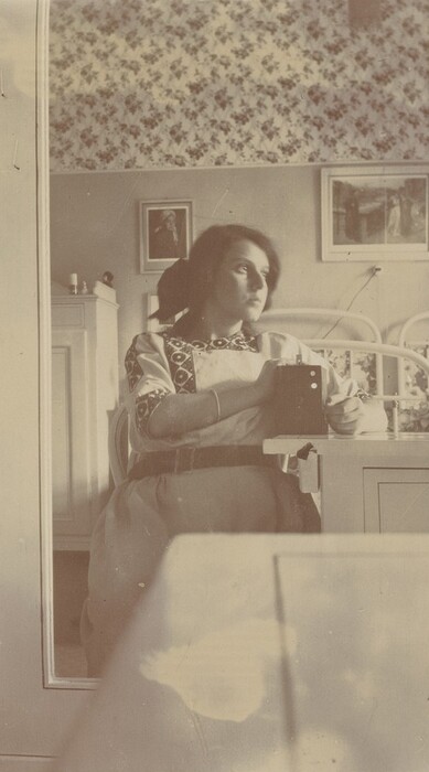 The image shows a woman from the waist up, seated and slightly angled to the left, looking toward the window. She has dark hair styled in a loose bun and wears a light-colored dress with a patterned design on the shoulder and sleeves. A belt cinches the dress at the waist, and she wears a bracelet on her left wrist. She holds a box camera in her right hand, resting it on a table. The background includes a well-lit room with floral wallpaper, framed pictures on the walls, a wooden cabinet, and a white wrought iron bed, suggesting a vintage decor theme.