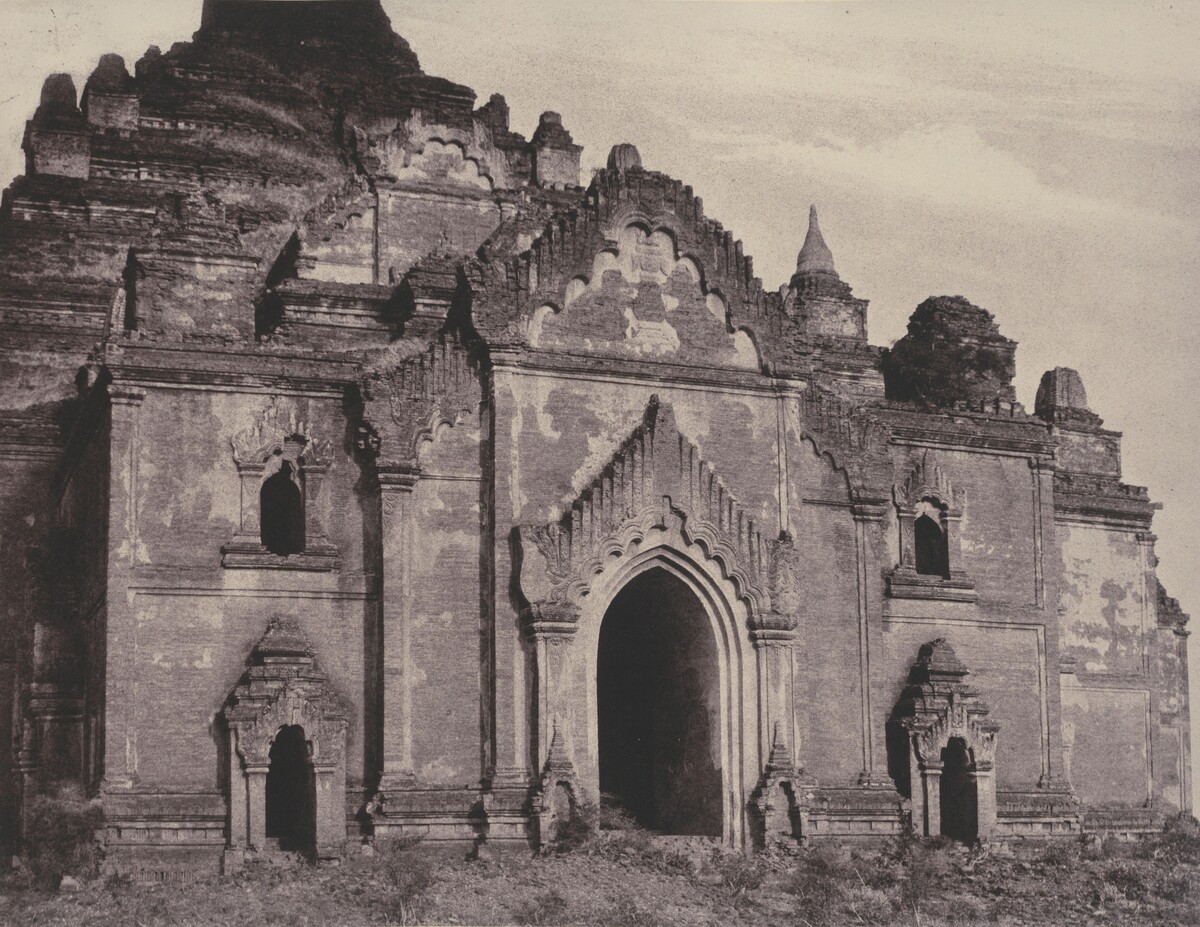 The image shows an ancient temple-like structure with intricate architectural details. The scene is closely cropped around the building, which features arched doorways, detailed carvings, and steps. The architecture is ornate, worn, and aged. The image is rendered in sepia tones, giving it a historical feel. The sky is relatively clear with some scattered clouds in the background.