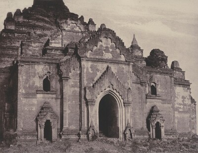 The image shows an ancient temple-like structure with intricate architectural details. The scene is closely cropped around the building, which features arched doorways, detailed carvings, and steps. The architecture is ornate, worn, and aged. The image is rendered in sepia tones, giving it a historical feel. The sky is relatively clear with some scattered clouds in the background.