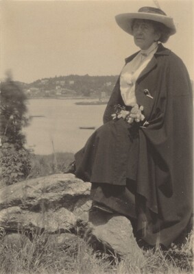 The image shows a woman sitting on a large rock outdoors. She is captured from the waist up, positioned in a three-quarters view. The woman has a mature appearance with soft facial features and short, dark hair mostly hidden under a wide-brimmed hat. She is wearing a dark cape over a light-colored blouse and holding a cluster of flowers around her midsection. The background includes a calm body of water with a distant shoreline populated with trees and sparse buildings.