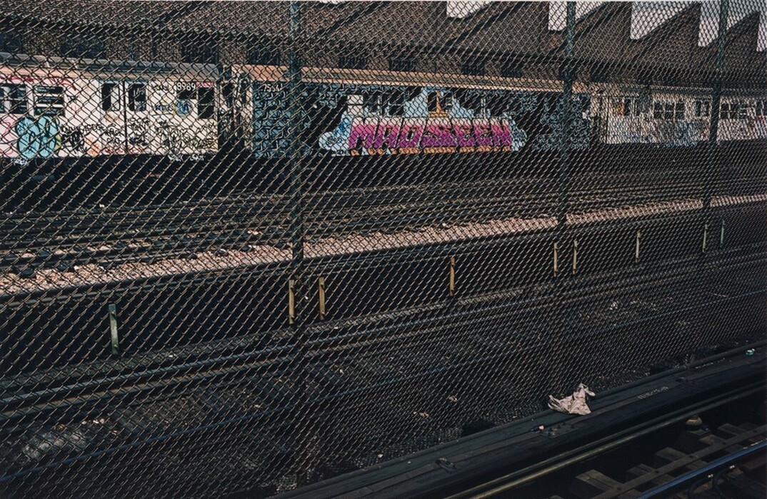 This is a photograph of a graffiti-covered train behind a chain-link fence. The image shows multiple train cars covered in colorful graffiti art in a train yard. The chain-link fence is prominent in the foreground, with a piece of discarded material on the ground nearby. The background includes an industrial building with a zigzag roof pattern.