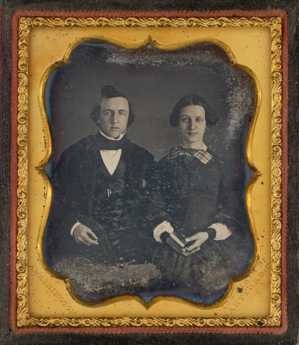 This grayscale photograph is a portrait of two people, visible from the knees up. They both have pale skin and dark hair. A man is seated on the left, facing forward. He has pale skin and short, dark hair styled into a curl on top of his head. He is wearing a dark suit with a white shirt and a black collar. A woman is seated on the right. Her hair is parted in the middle and styled into bunches above her ears. She is wearing a dark dress with a checkered pattern around the collar and white cuffs. She holds a small book in her hands. The backdrop is plain and dark. The photograph is enclosed in a gold frame with some dark specks and discolorations. Around that frame is a dark wooden frame with some red trim.