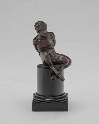The sculpture portrays a satyr, a mythological creature with a human upper body and a goat-like lower body. The satyr is depicted seated with its arms bound behind its back, legs crossed, and head slightly tilted. The bronze sculpture showcases detailed musculature, facial features, and textured hair. It is placed on a black stone pedestal.