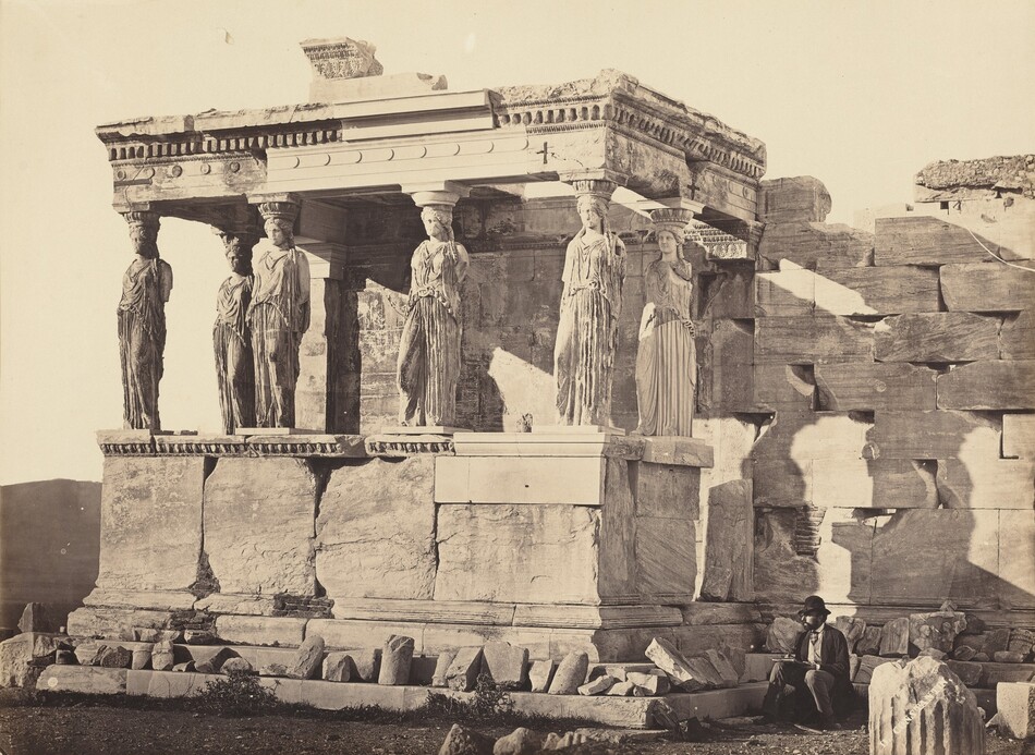 The remains of a rectangular porch-like portico are photographed in tones of ash-brown and black in this horizontal albumen print. The roof of the portico is held up by six marble columns carved into the shape of women wearing robes that fall in long, vertical folds. The women all face forward, and some of their features are weathered. All are missing their arms. The portico and structure behind it are constructed with massive, squared blocks. A man wearing a suit and a bowler hat sits drawing in the corner where the portico meets the bulk of the building, near the lower right corner of the photograph. Fragments of reliefs and other architectural ornaments are lined up behind and next to him. A few scrubby plants grow on the dirt ground, and the sky above is a washed out white. White text angled up across the lower right corner reads, “D. CONSTANTIN.”