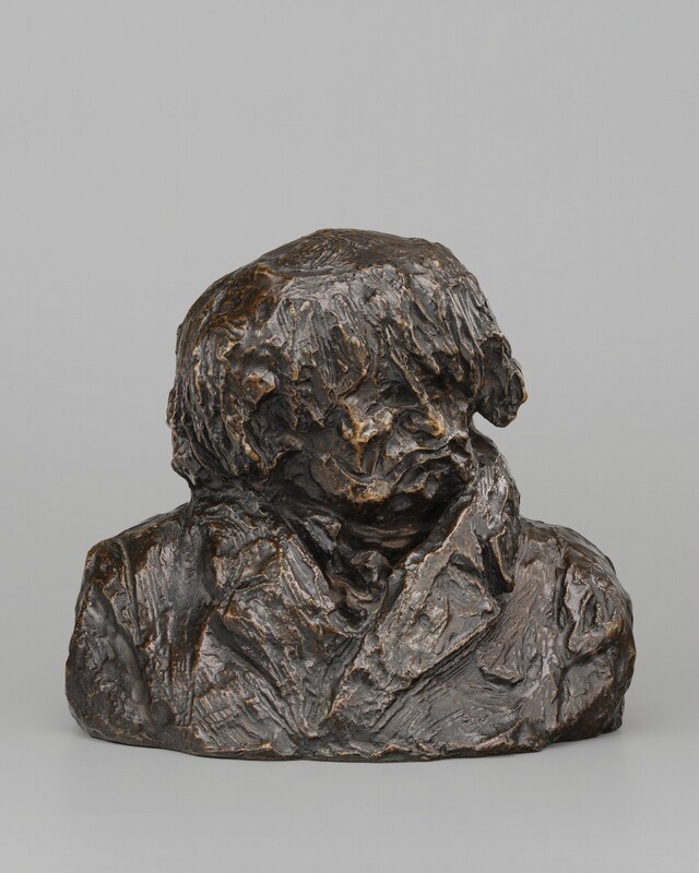 At first glance, this bronze sculpture resembles a lump of clay loosely formed into the head and shoulders of a man. In this photograph, his shoulders are squared toward us as he looks to our right. His hair is pushed forward in rope-like spikes over deep-set eye sockets. He has a sharp, thin nose, and his lips purse in an exaggerated scowl. He seems to wear a cravat nestled into the high lapels of his coat. The surface of the bronze is mostly brown except where the edges and ridges are gold, as if having been burnished through handling.