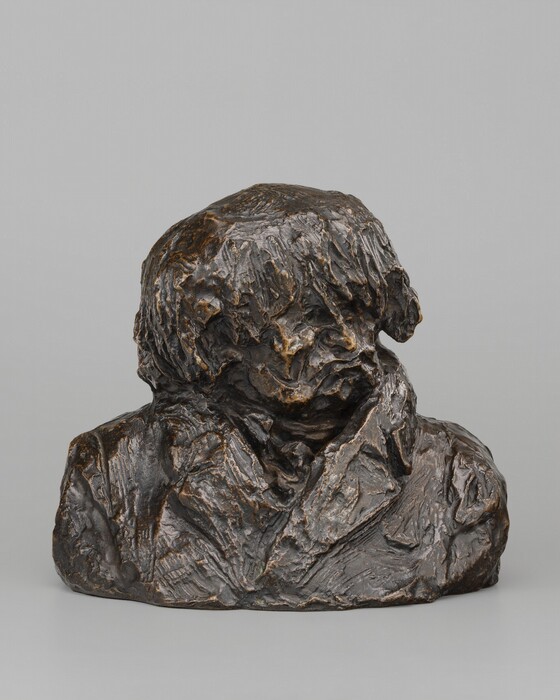 At first glance, this bronze sculpture resembles a lump of clay loosely formed into the head and shoulders of a man. In this photograph, his shoulders are squared toward us as he looks to our right. His hair is pushed forward in rope-like spikes over deep-set eye sockets. He has a sharp, thin nose, and his lips purse in an exaggerated scowl. He seems to wear a cravat nestled into the high lapels of his coat. The surface of the bronze is mostly brown except where the edges and ridges are gold, as if having been burnished through handling.
