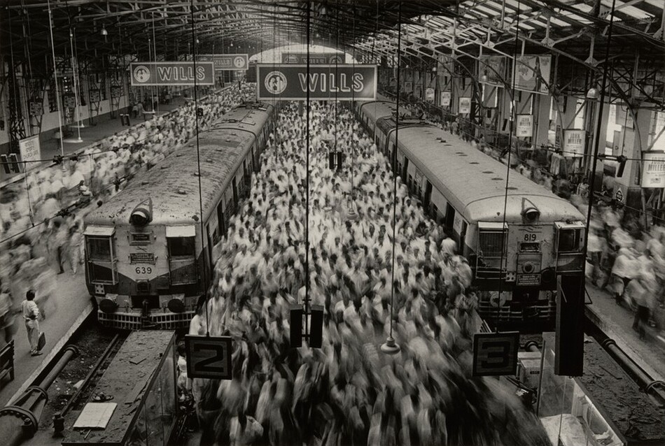 This is a photograph of a train station with two trains on platforms and a crowd of moving people. Two trains are parked on platforms labeled 2 and 3. Above the platforms are advertisements for 'WILLS' and there are structures supporting the roof. The image shows a dynamic scene with blurry movements of people in the station. There are industrial elements like metal beams, large windows, and overhead lights. The crowd adds a sense of busy activity as people move around the station.