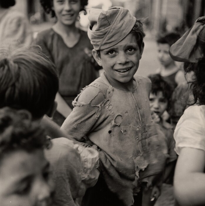 The image shows a group of children with a focus on a young boy who is captured from the waist up. He is facing the camera with a slight tilt to his body. His facial features include big, bright eyes, a wide smile revealing his teeth, and dirt on his face. His hair is covered by a makeshift cap or cloth. The boy is wearing a worn and tattered shirt with multiple holes and frayed edges. Around him are other children, some partially visible and slightly out of focus, standing closely together in an outdoor, urban environment.