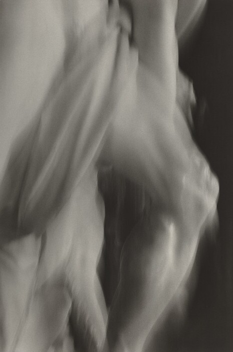 The image shows soft, flowing shapes and blurred lines creating an abstract form. The arrangement of these lines and shapes suggests fluid motion. The interplay of light and shadow adds depth, resembling folds of draped cloth or contours of a moving figure.