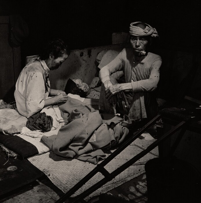 This is a photograph of a dimly lit interior scene featuring rustic materials and textiles. The image captures a setting where two figures are in a room or enclosure, surrounded by several layers of thick fabric, cushions, and possibly other bedding materials. The lighting is dramatic, casting deep shadows and highlighting the texture of the fabrics and the wooden surfaces around. The ambiance suggests an intimate, perhaps domestic environment with a sense of seclusion or focus on rest and contemplation. The black and white palette enhances the contrast, bringing attention to the various textures and subtle details within the setting.