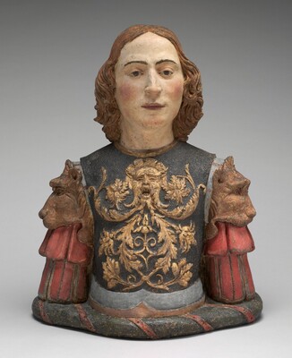 The sculpture depicts a bust of a person with a serene expression and shoulder-length wavy hair. The character is adorned with intricately detailed armor, which features a central motif of a grotesque face surrounded by floral patterns in gold. The armor is primarily a dark, muted hue, accented with red sleeves that resemble fabric folds, and two horse-like figures on the shoulders. The sculpture is created from what appears to be a painted, textured material, providing a realistic yet stylized representation of a historical or mythological figure. The face contrasts with the darker tones of the armor, enhancing the lifelike appearance of the sculpture.